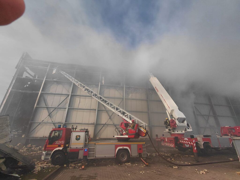 Strażacy gaszą pożar w hali przemysłowej, używając wozu strażackiego z drabiną i armatki wodnej. W tle widoczny dym.