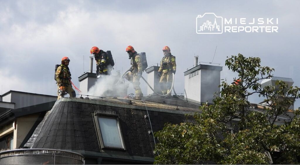 Czterej strażacy w hełmach gaszą dymiący dach budynku, używając węża strażackiego. W tle widać drzewa i chmurki.