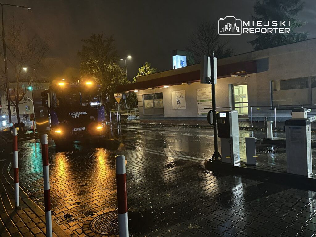 Ciężarówka Scania z włączonymi światłami awaryjnymi stoi na mokrej nawierzchni przy budynku, w nocy.