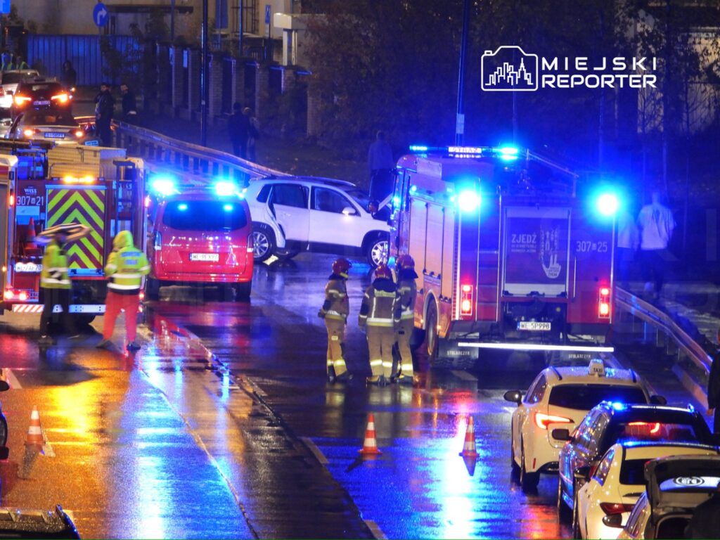 Strażacy w żółtych i czerwonych uniformach pracują przy wypadku drogowym na mokrej jezdni, obok zaparkowanych samochodów.