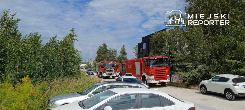 Dwa czerwone wozy strażackie stoją na wąskiej drodze otoczonej drzewami, obok zaparkowane samochody osobowe.