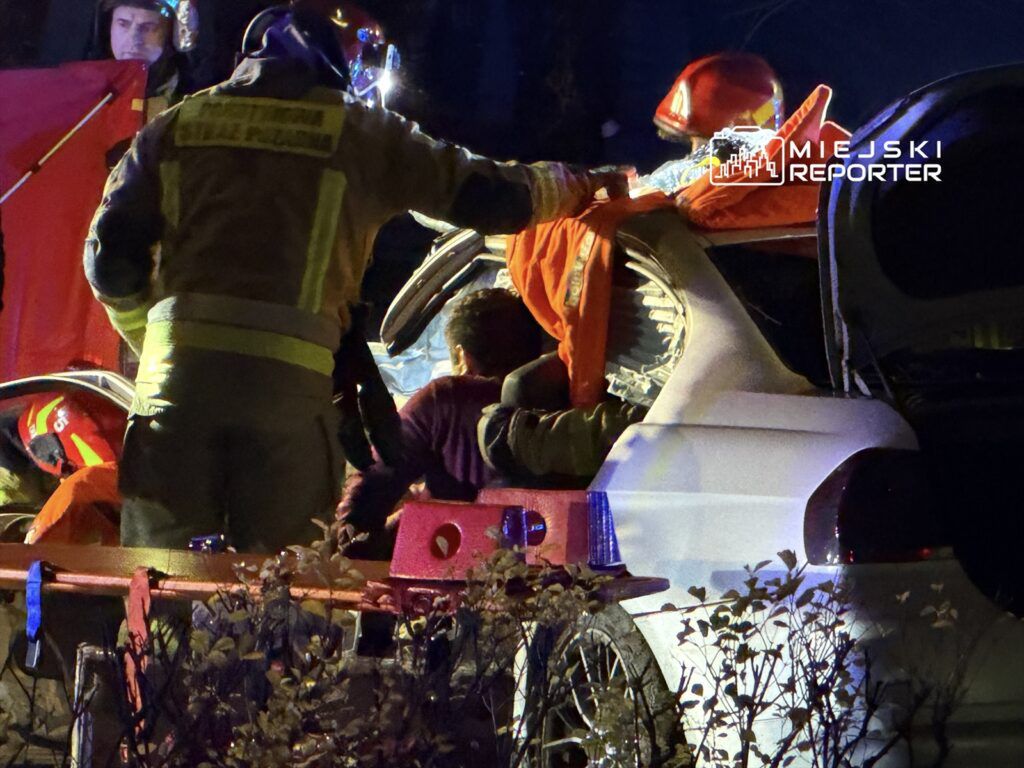 Strażacy udzielają pomocy osobie poszkodowanej w rozbitym samochodzie w nocy, otoczeni przez pojazdy ratunkowe.