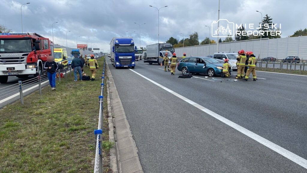 Grupa strażaków udziela pomocy poszkodowanemu w wypadku drogowym na autostradzie, obok uszkodzonego samochodu.