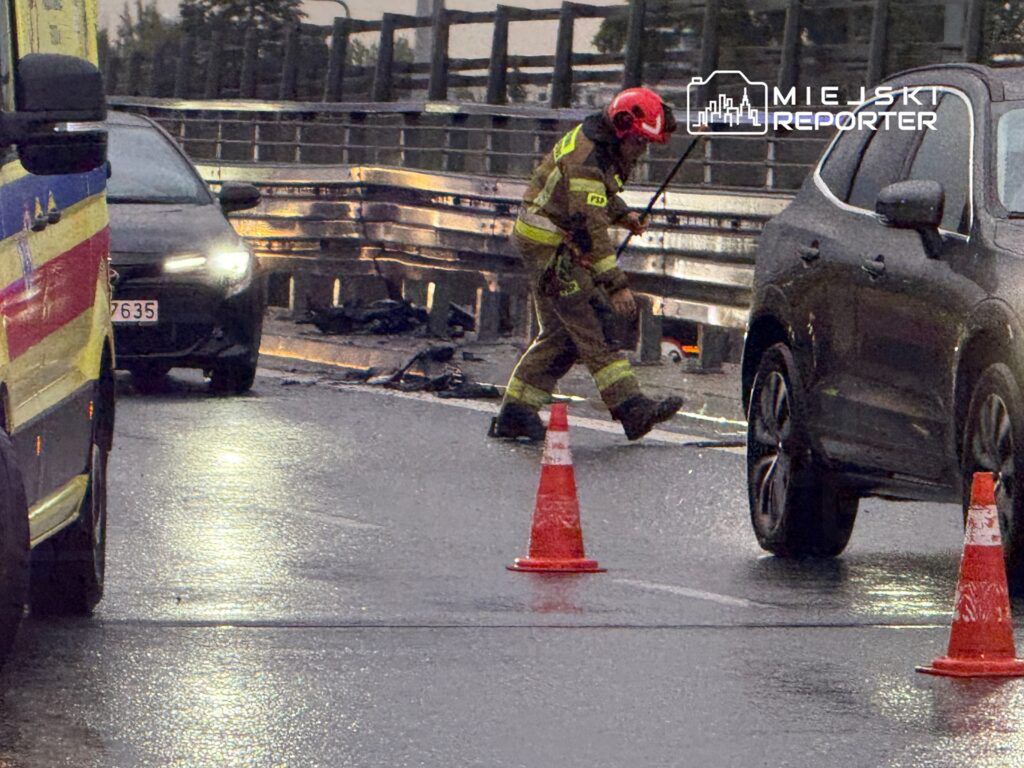 Strażak w kasku i mundurze bada miejsce zdarzenia na mokrej jezdni, obok oznakowanie w postaci pachołków.