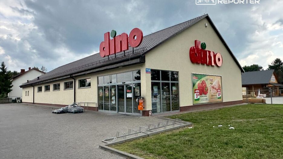 Supermarket „Dino” z dużym, kolorowym logo, z przeszklonymi drzwiami i reklamami na elewacji, w otoczeniu parkingu.