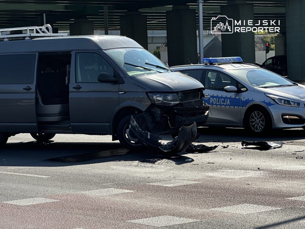 Uszkodzony szary van z otwartymi drzwiami stoi na drodze, obok niego policyjny radiowóz z napisem "POLICJA".