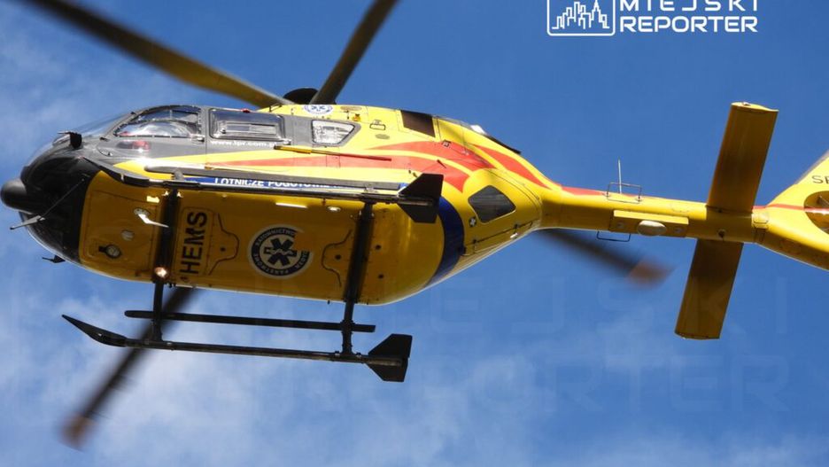 Żółty śmigłowiec ratunkowy HEMS przelatuje nad miastem, wirniki kręcą się w błękitnym niebie.