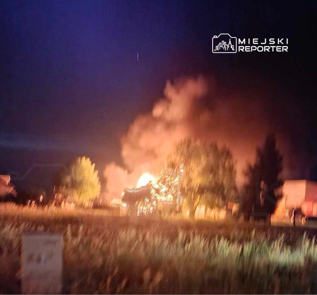 Pożar budynku emitującego gęsty dym i płomienie w nocnej scenerii, otoczony przez drzewa i trawę.