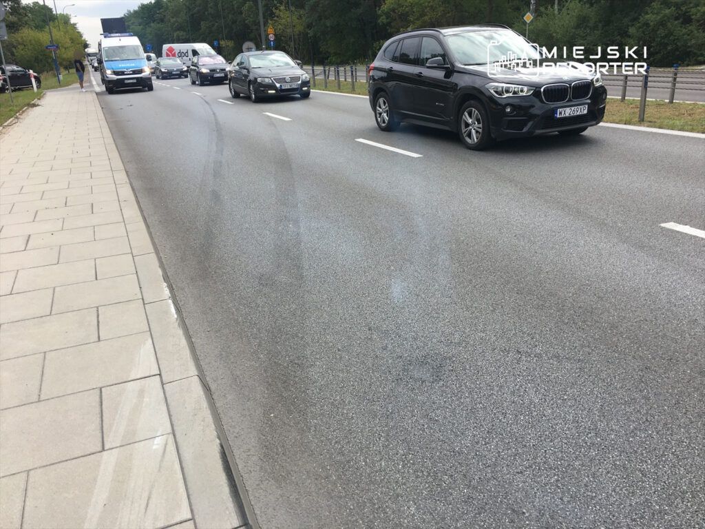 Ruch samochodowy na jezdni, obok chodnika z płytami, w otoczeniu drzew i znaków drogowych.
