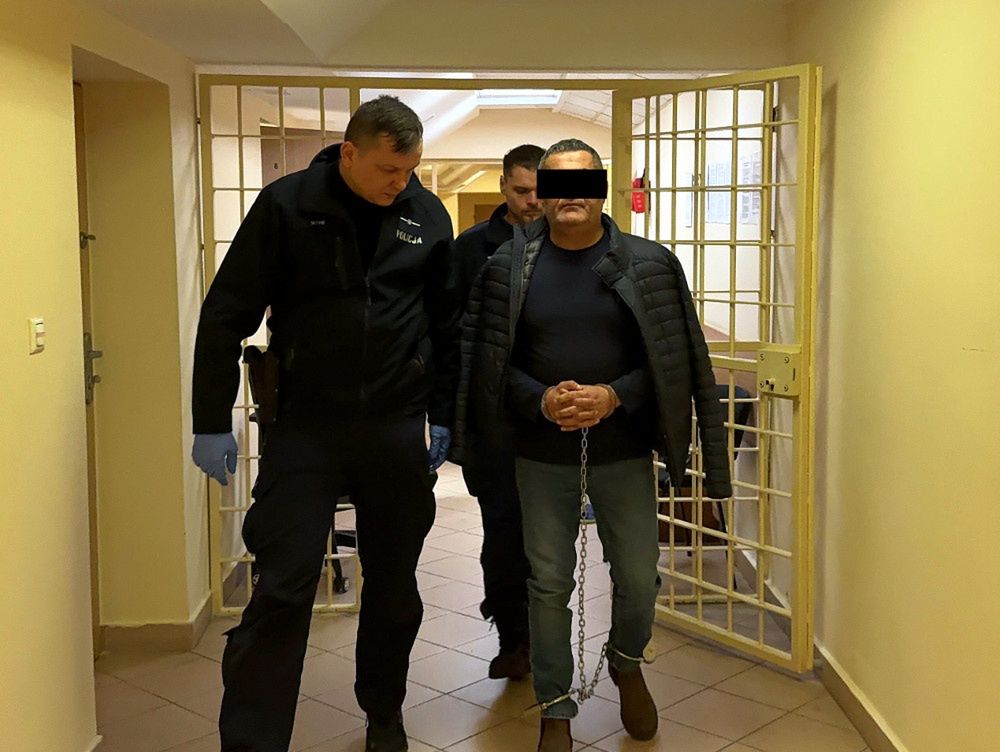 Mężczyzna w kajdankach prowadzony przez policjantów w korytarzu budynku.