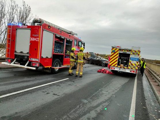 Strażacy w hełmach pracują przy wypadku drogowym na moście, obok unieruchomionego samochodu osobowego.