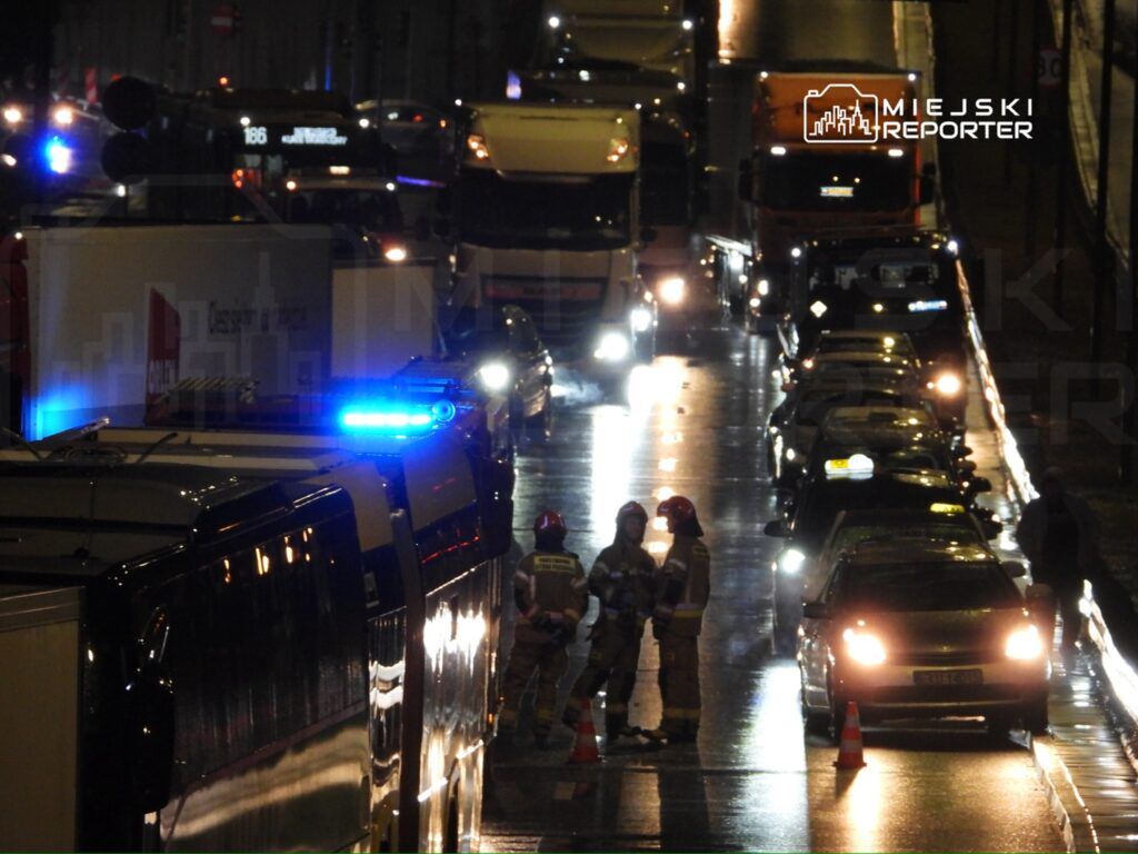 Ratownicy w mundurach rozmawiają na mokrej ulicy, obok zaparkowanych samochodów, w tle widać pojazdy z włączonymi sygnałami.