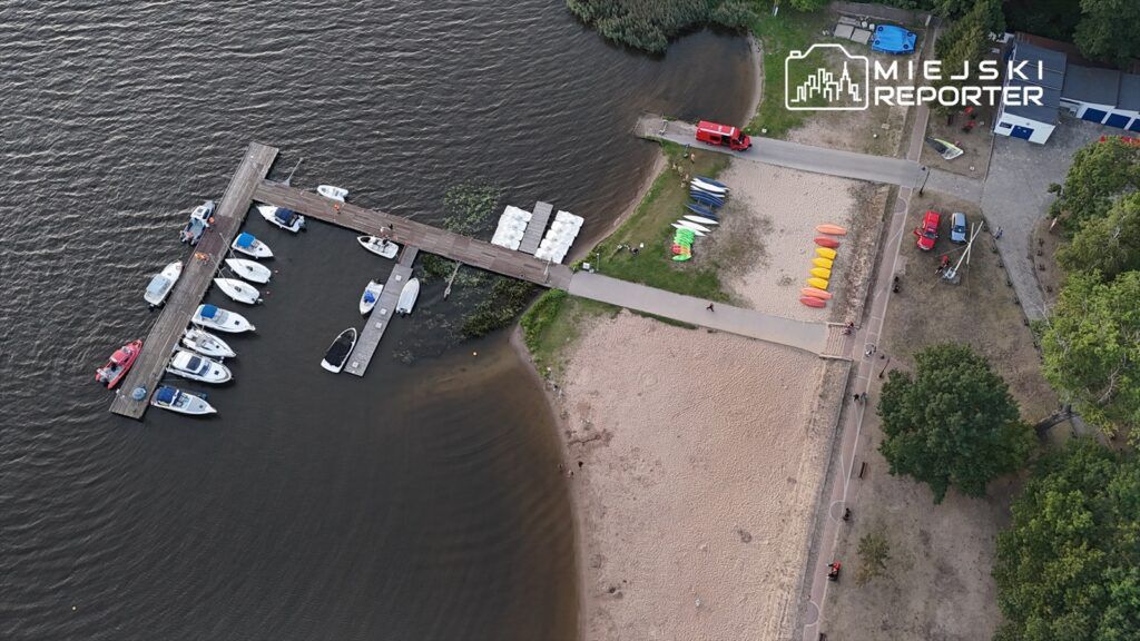 Molo z łodziami przy brzegu jeziora, obok plaża z leżakami i sprzętem wodnym, w tle zieleń drzew.