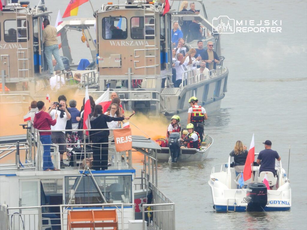 Grupa osób z flagami na pokładzie statku obserwuje akcję ratunkową na wodzie, gdzie zespół ratowników wchodzi do łodzi.