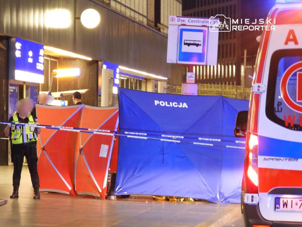 Policjant zabezpiecza miejsce zdarzenia przy dworcu, z niebieskimi i pomarańczowymi namiotami oraz karetką w tle.
