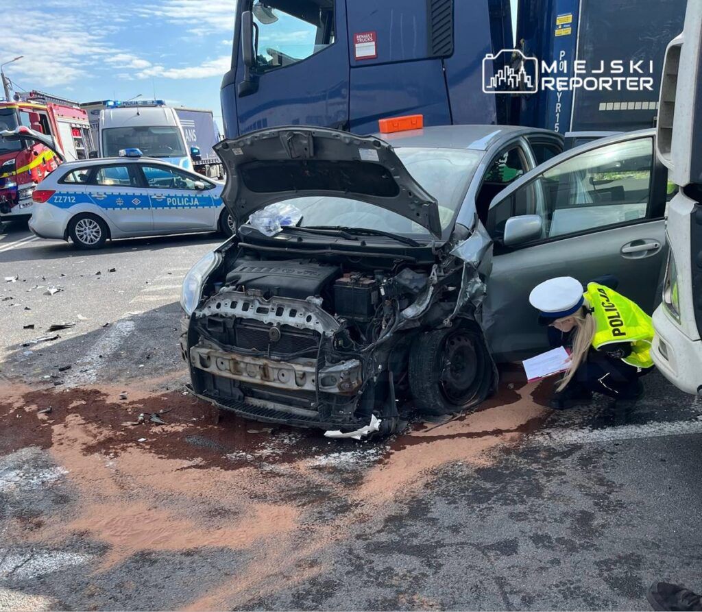 Kobieta w mundurze policji bada wrak samochodu osobowego na drodze, obok stoją ciężarówki i pojazd policyjny.