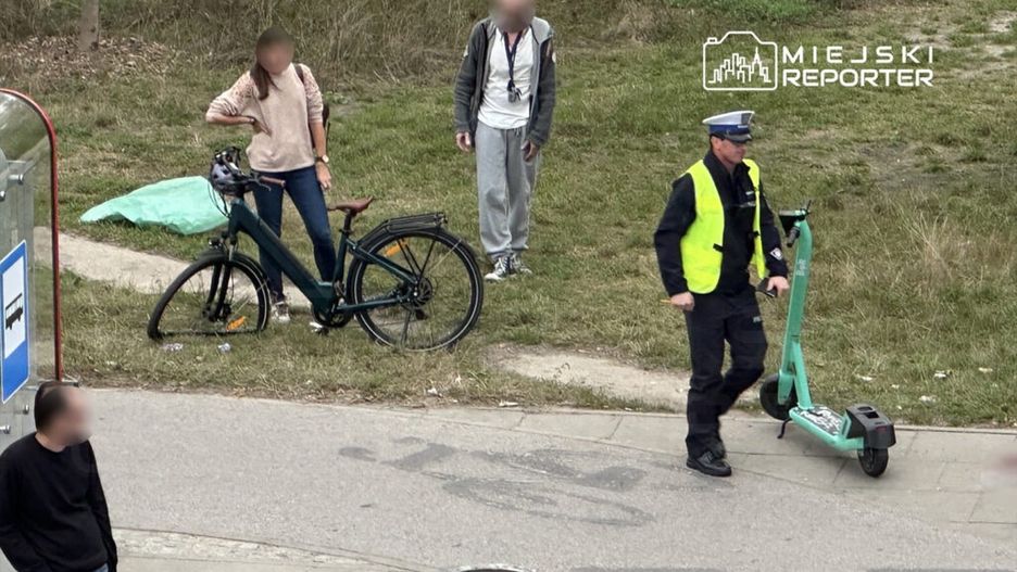 Mężczyzna w mundurze policyjnym prowadzi elektryczną hulajnogę w parku, obok stoją rower i grupa osób.
