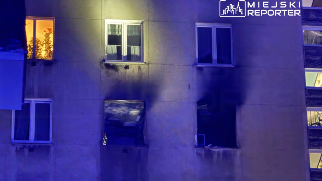 Na elewacji budynku widać okna, z których jedno jest oświetlone, a obok znajduje się okno z uszkodzeniami po pożarze.