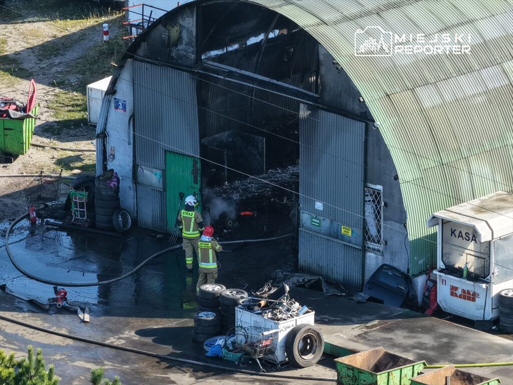 Strażacy gaszą pożar w zniszczonym warsztacie, obok leżą opony i odpady metalowe. W tle widoczny budynek z kasą.