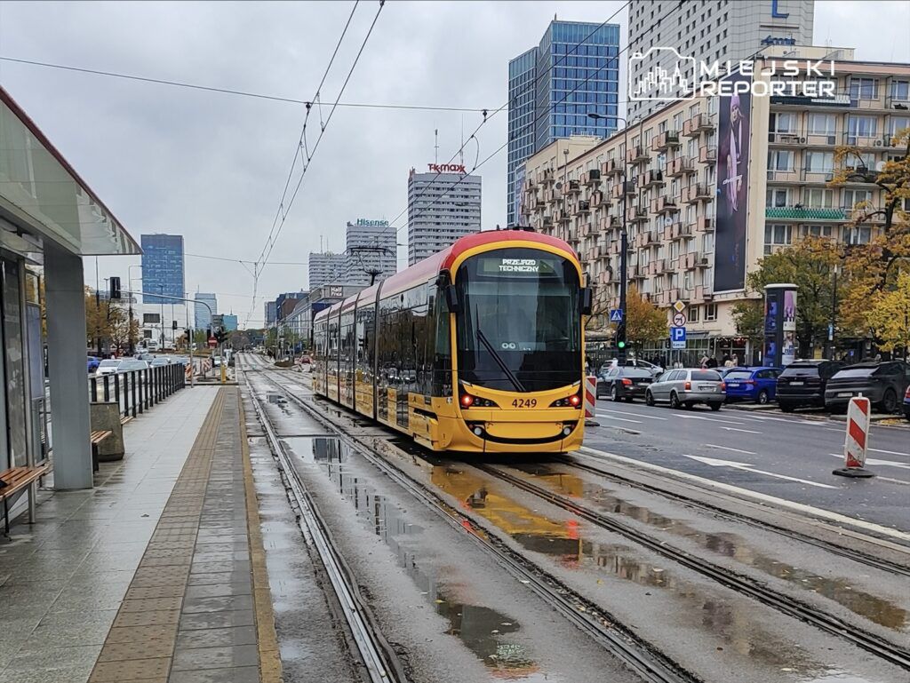 Tramwaj w kolorze żółtym przemieszcza się po mokrej jezdni w miejskim otoczeniu, otoczony nowoczesnymi budynkami.