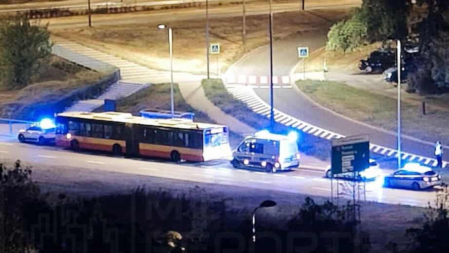 Autobus z włączonymi światłami awaryjnymi zatrzymany na drodze, otoczony przez radiowozy policyjne z niebieskimi sygnałami świetlnymi.