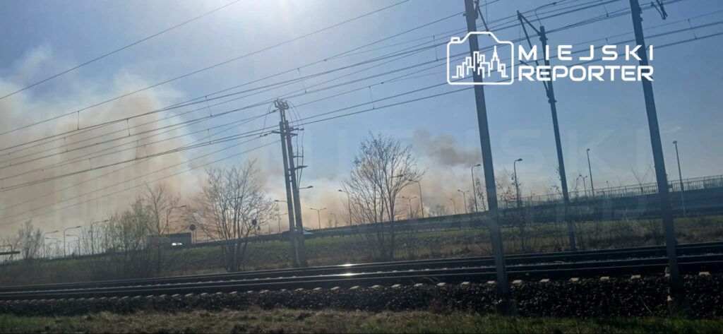 Gęsty dym unoszący się nad terenami przemysłowymi w pobliżu torów kolejowych w słoneczny dzień.