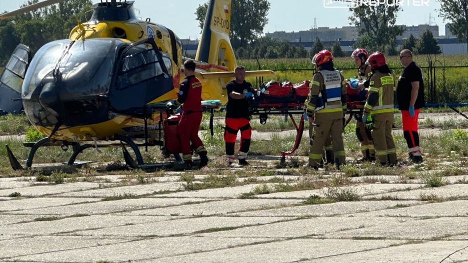 Ratownicy medyczni i strażacy pracują przy helikopterze, przygotowując nosze do transportu poszkodowanego.
