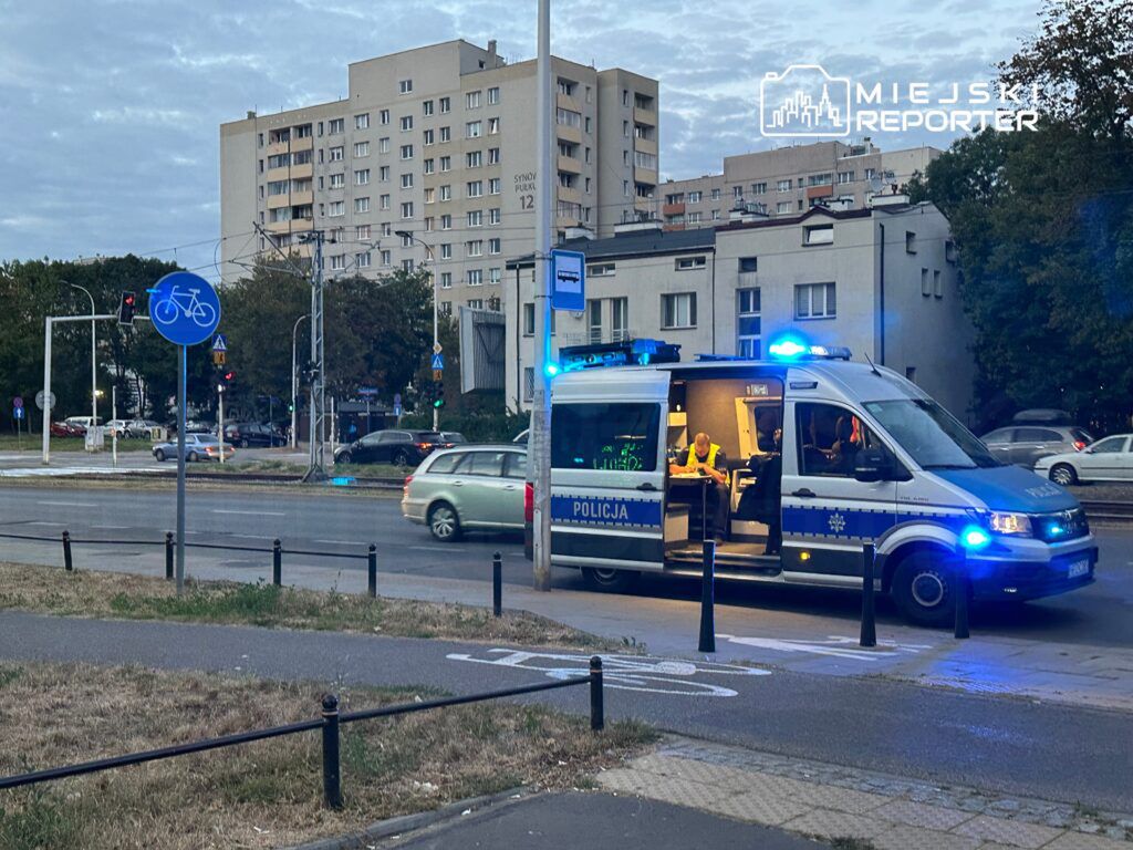 Policyjny radiowóz z włączonymi sygnałami świetlnymi zatrzymany przy ulicy, obok znaku drogowym dla rowerzystów.