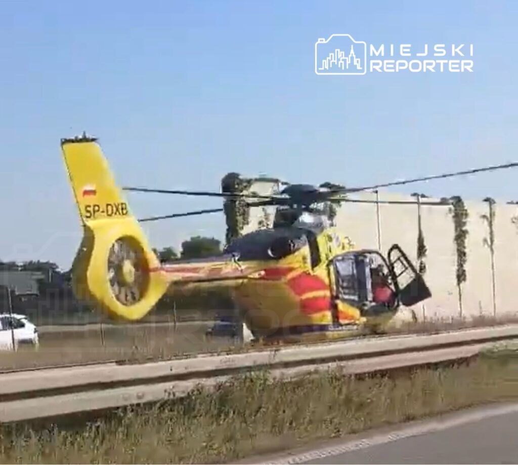 Śmigłowiec ratunkowy lądujący obok drogi, z otwartymi drzwiami i widocznym pilotem.