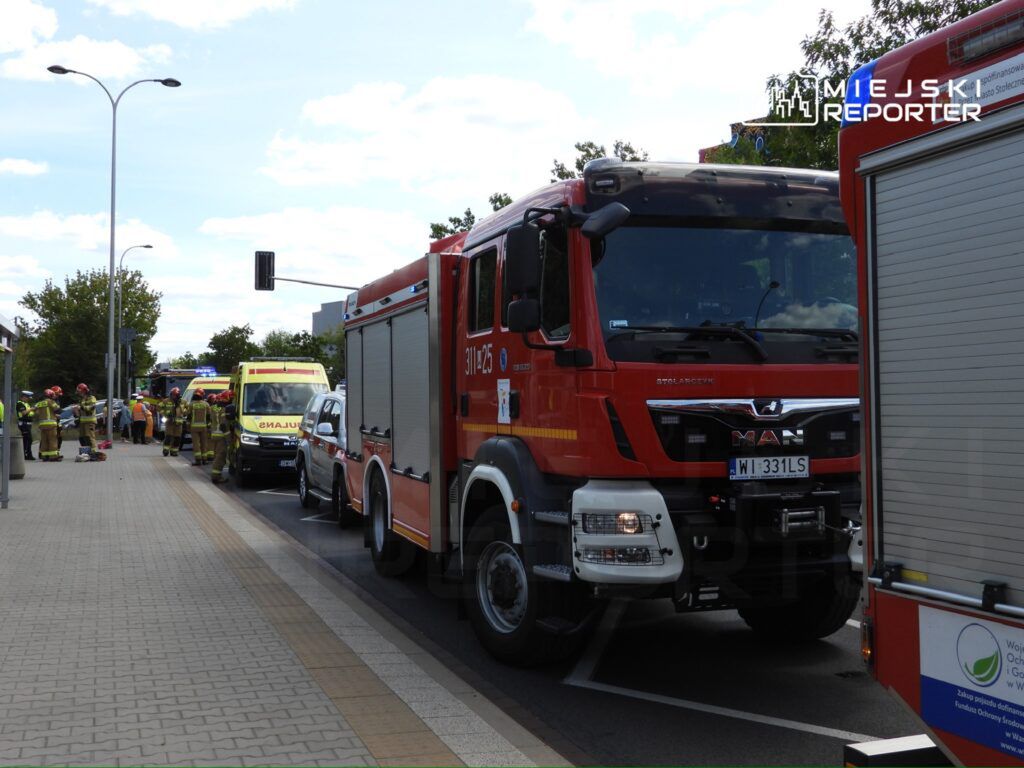 Strażacy w mundurach pracują przy wozach strażackich i karetce na miejskiej ulicy, w otoczeniu zieleni.