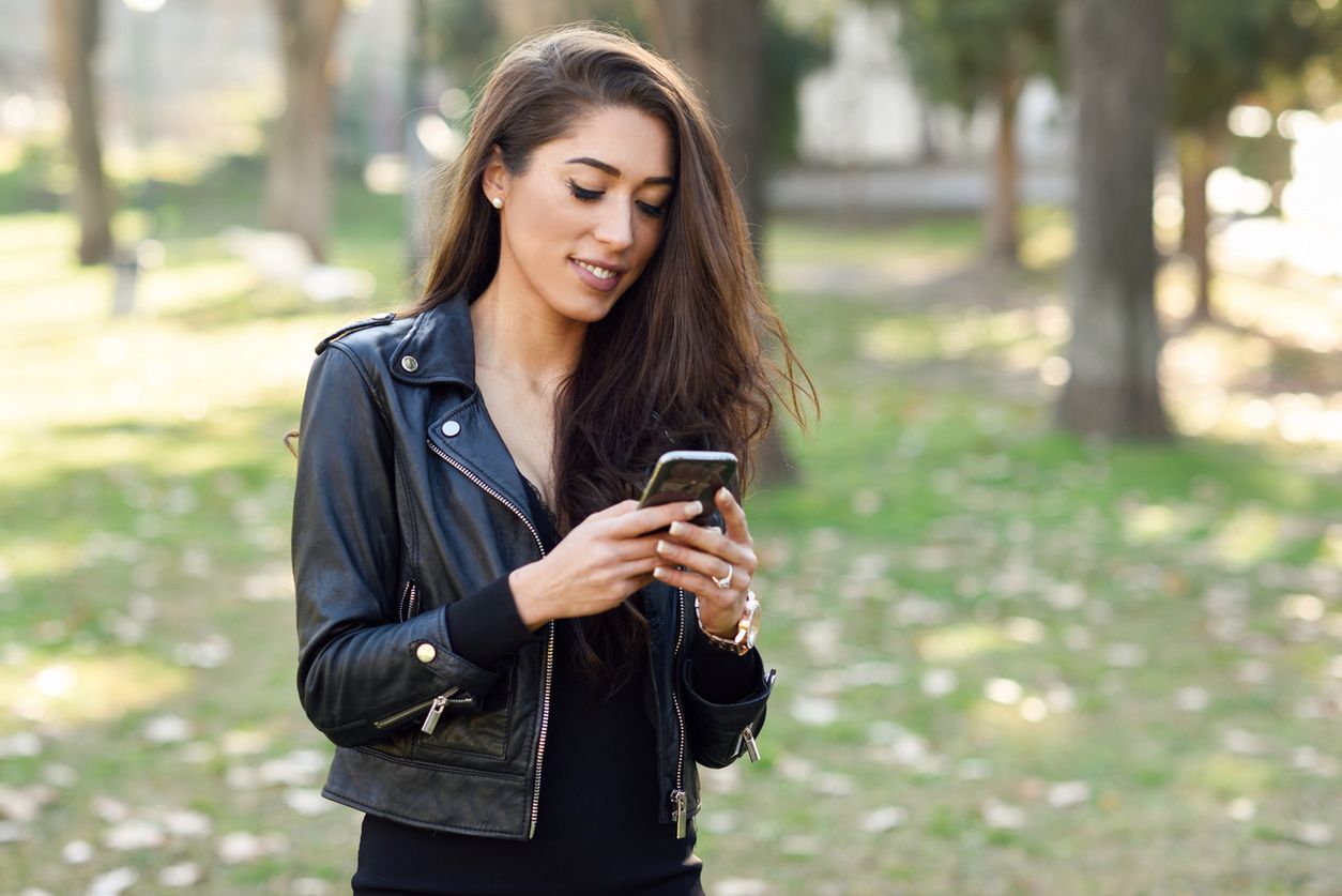 Young woman using a smartphone in an urban park. Girl wearing leather jacket. 