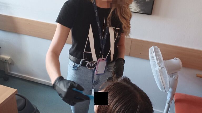 Kobieta w czarnej koszulce zakłada rękawice i wykonuje czynność na głowie siedzącej osoby w pomieszczeniu biurowym.