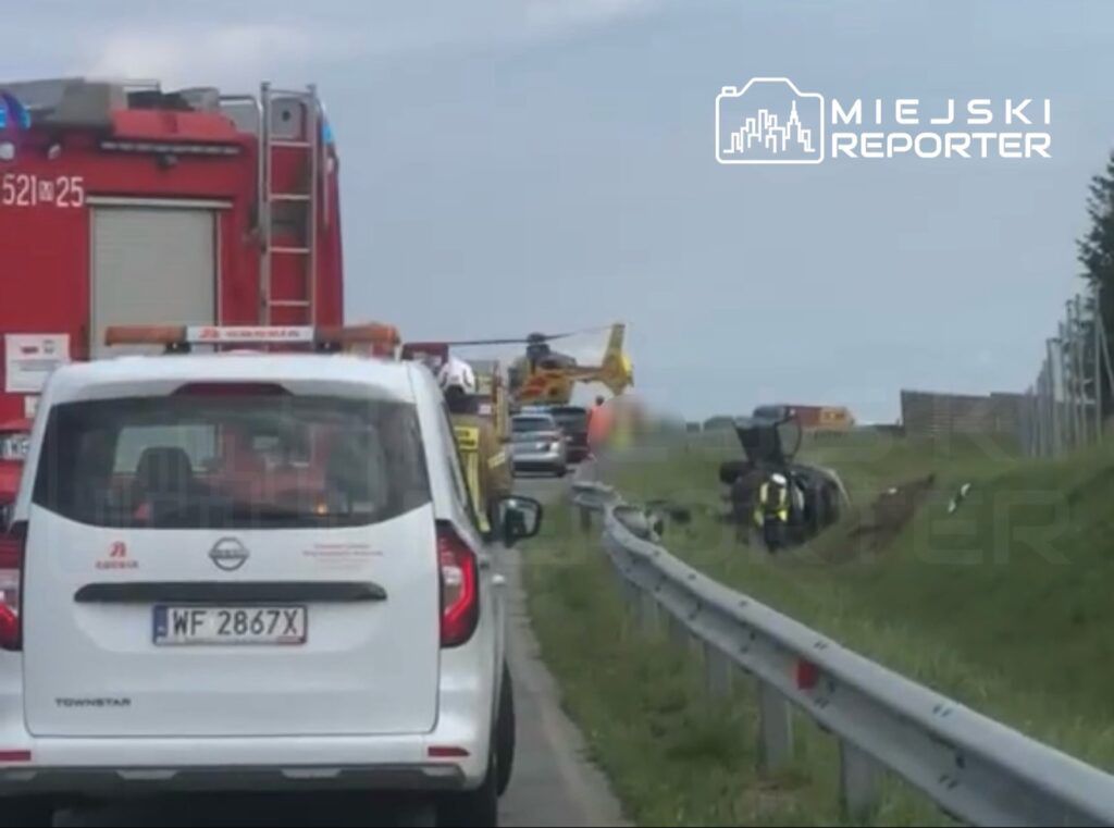 Na drodze stoi kilka pojazdów, w tym samochód strażacki, obok lądował śmigłowiec ratunkowy, a na poboczu leży przewrócony motocykl.