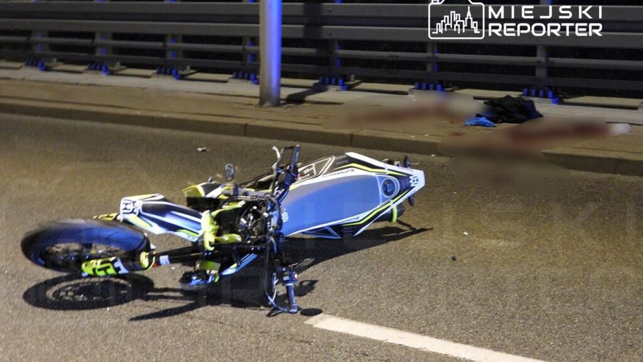 Motocykl leżący na jezdni, obok widoczne plamy krwi oraz ciuchy. Nocne oświetlenie ulicy.