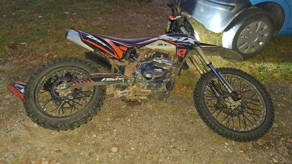 Motocykl terenowy o pomarańczowo-czarnym malowaniu stoi na ziemnym podłożu obok niebieskiego samochodu.