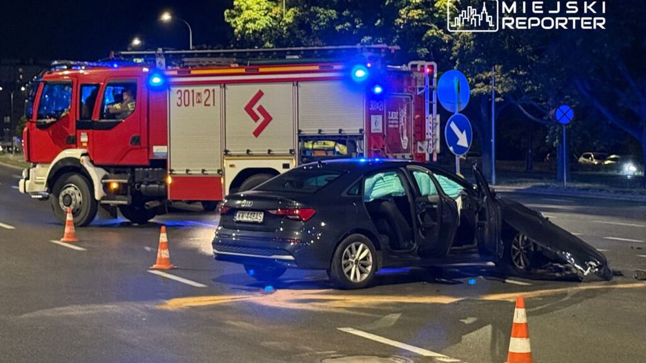 Czarny samochód osobowy z otwartymi drzwiami stoi na jezdni po wypadku, obok czerwony wóz strażacki z włączonymi sygnalizatorami.