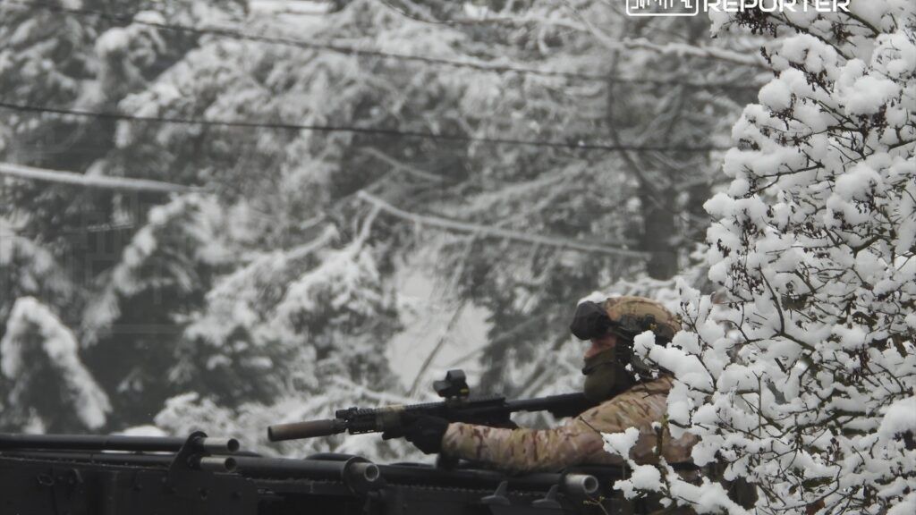 Żołnierz w kamuflażu strzeże terenu z pojazdu opancerzonego, otoczonego zaśnieżonymi drzewami.