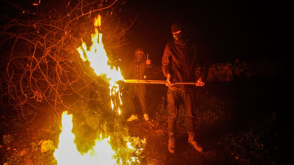 NABLUS, WEST BANK, PALESTINE - 2023/03/01: Masked Palestinians carrying wooden sticks stand around a fire on guard during the night to fend off attacks by the Jewish settlers in south of Nablus in the West Bank. The Settler attacks on Palestinians have increased, especially in the south of Nablus. In the town of Hawara, settlers damaged and burned dozens of homes, shops, and cars. Palestinian Prime Minister Muhammad Shtayyeh said that the total losses were estimated at nearly $5 million. (Photo by Nasser Ishtayeh/SOPA Images/LightRocket via Getty Images)
