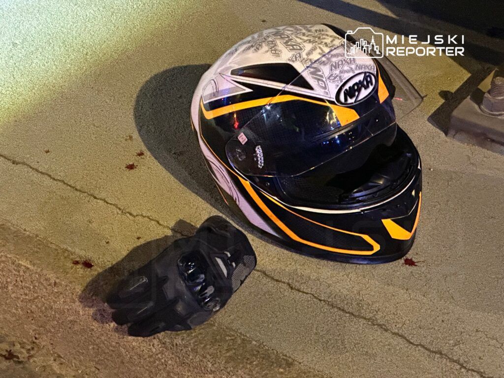 Kask motocyklowy z widocznymi śladami krwi leży na asfalcie obok rękawicy motocyklowej.