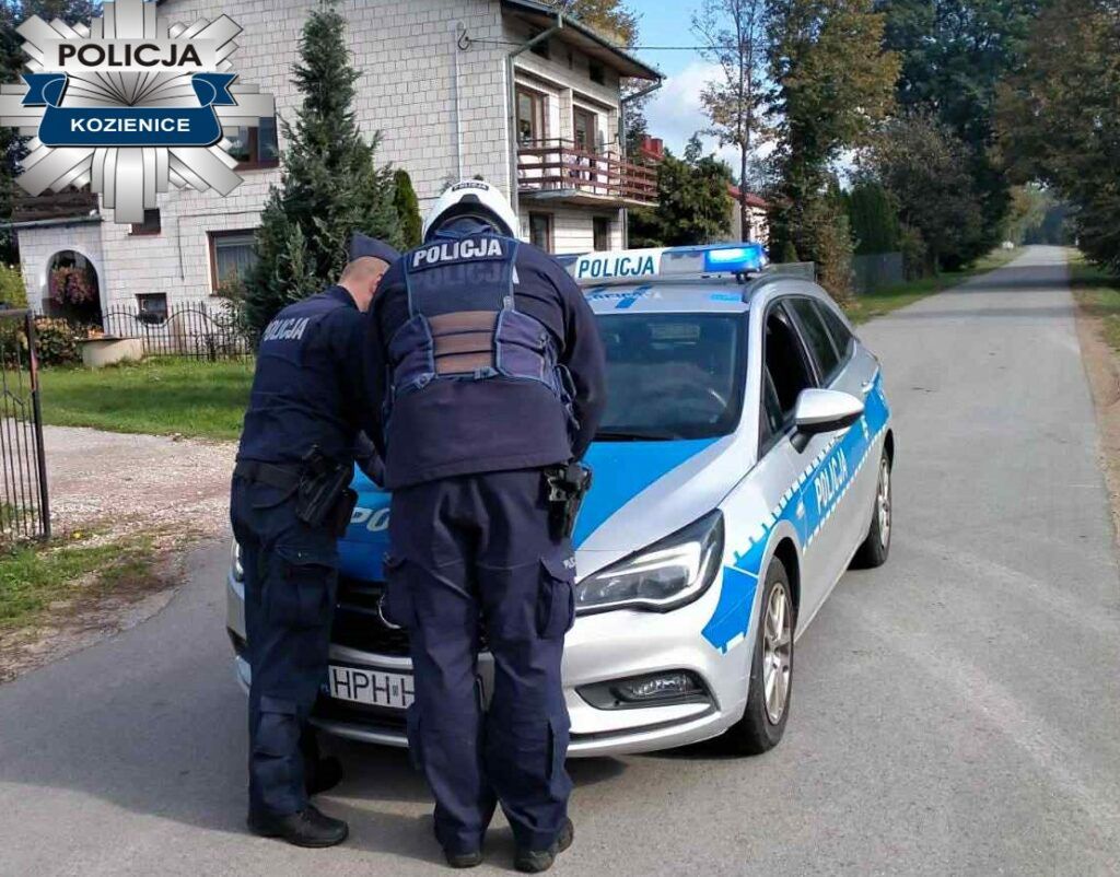 Dwaj policjanci w mundurach przeszukują samochód służbowy na wiejskiej drodze, obok domów jednorodzinnych.