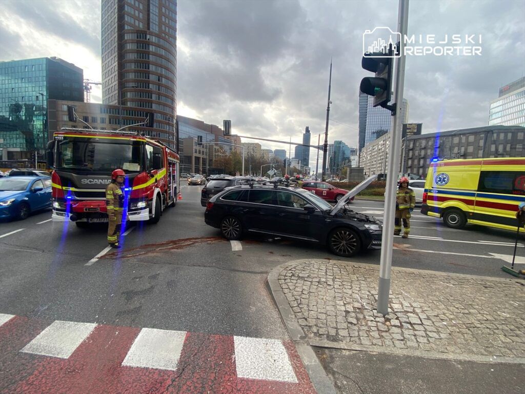 Czterech strażaków pracuje przy unieruchomionym samochodzie osobowym na ruchliwej ulicy w mieście. W tle widoczna karetka.