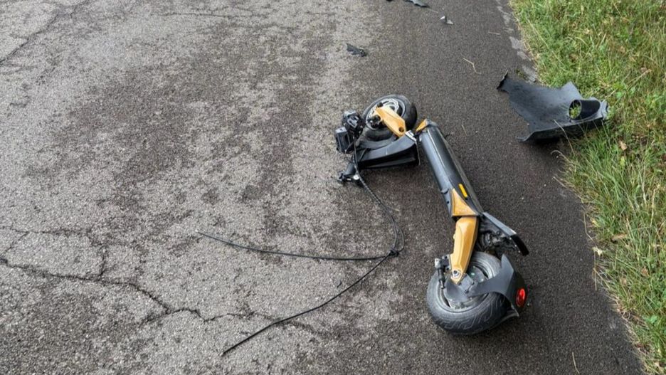 Złamana hulajnoga leży na asfaltowej drodze, obok widoczne fragmenty plastikowych osłon.