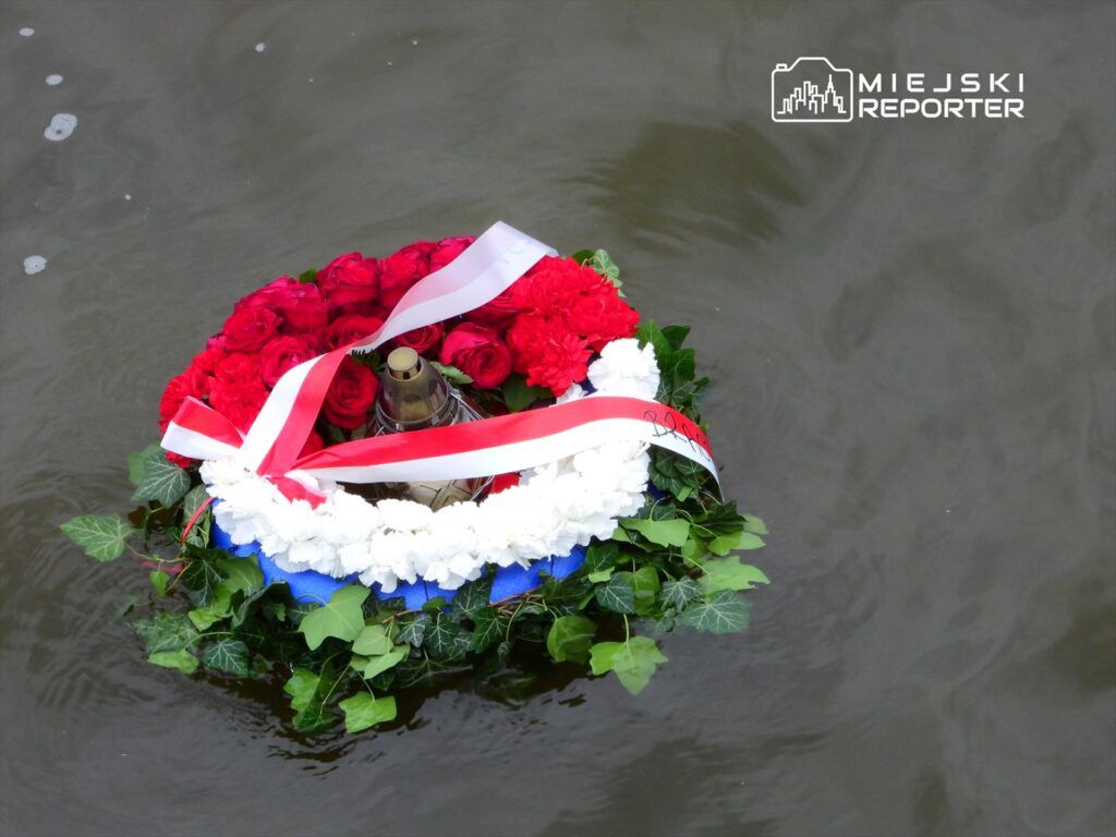 Wieniec z czerwonych róż i białych goździków z metalowym zniczem unosi się na wodzie, otoczony zielonymi liśćmi.