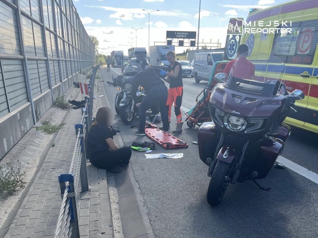 Grupa osób udziela pomocy poszkodowanemu motocykliście na ruchliwej drodze, obok karetki pogotowia.