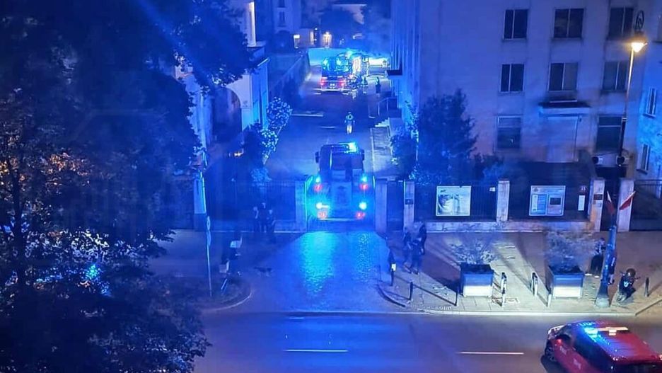 W nocy na ulicy widoczne niebieskie światła radiowozów i wozów strażackich, w tle budynki mieszkalne.
