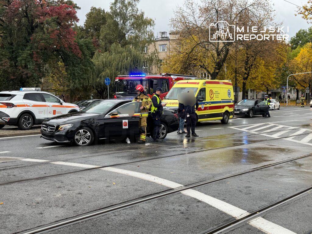 Czarny samochód osobowy stoi na ulicy, obok grupy ratowników oraz policjantów, w tle widać pojazd straży pożarnej.