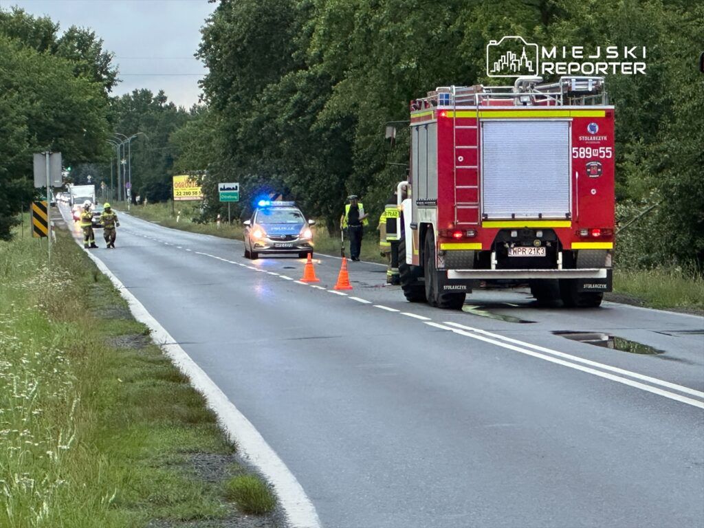 Strażacy i policjanci zabezpieczają miejsce wypadku na drodze, podczas gdy samochód strażacki stoi w tle.