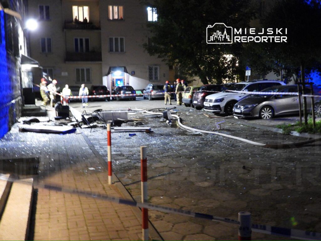 Grupa strażaków zabezpiecza miejsce zdarzenia, gdzie widać zniszczone mienie i szkło na chodniku.