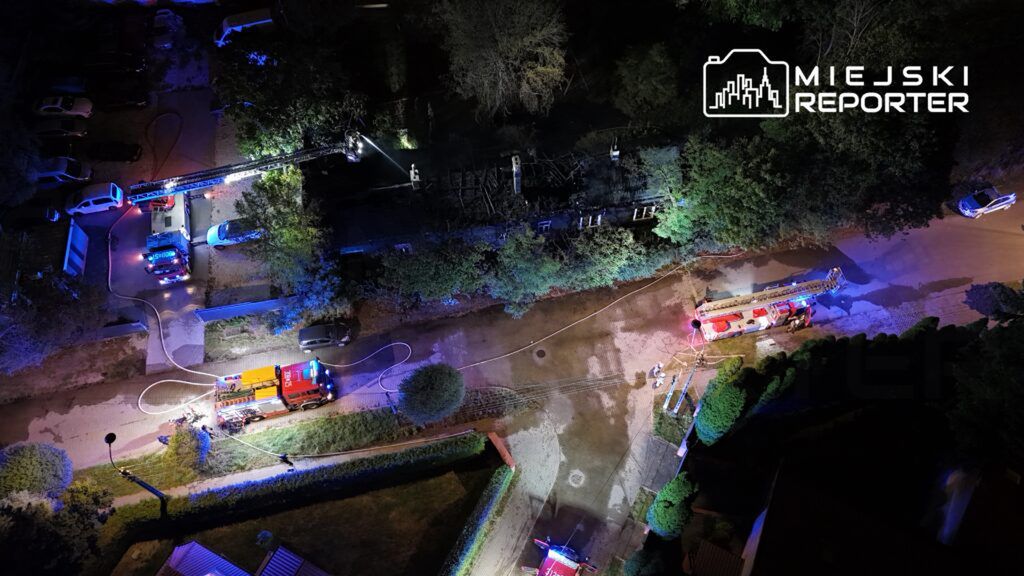 W nocy strażacy gaszą pożar w budynku, używając wozów strażackich oraz drabiny, na oświetlonym osiedlu.