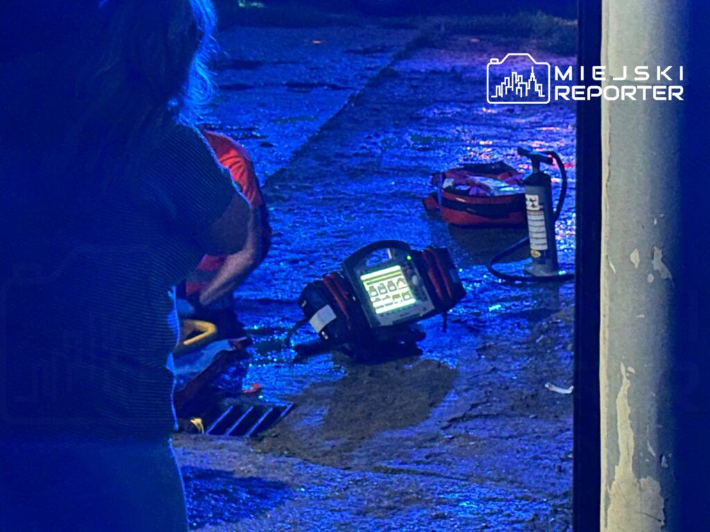 Kobieta w koszulce w paski obserwuje pracę ratowników w niebieskim świetle, obok leży sprzęt ratunkowy.